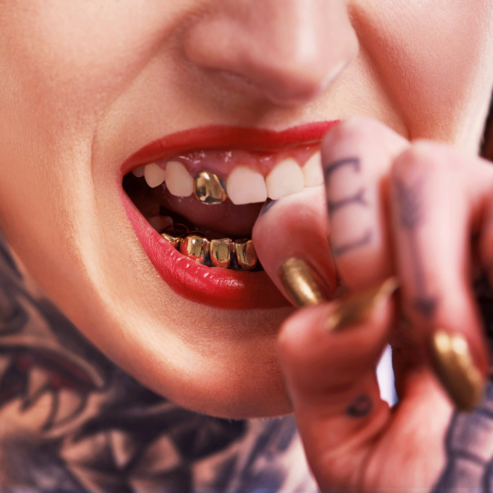 Close-up of a person showing off gold grillz and tattooed hands, highlighting luxury fashion in hip-hop culture.
